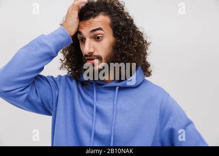 Bild eines verärgerten unshaven Mannes in legeren Sweatshirt, das aufschäumt und seinen Kopf isoliert über weißem Hintergrund ergreift Stockfoto