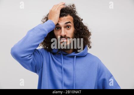 Bild eines verwirrten, unrasierten Mannes in lässigem Sweatshirt, das aufschäumt und seinen Kopf isoliert über weißem Hintergrund ergreift Stockfoto