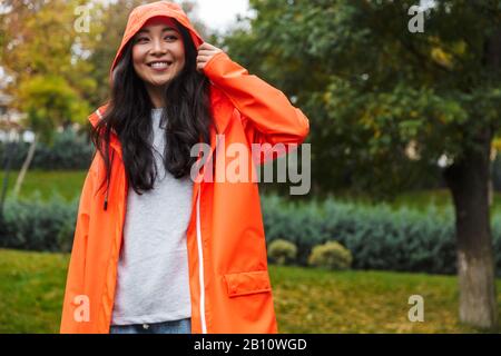 Lächelnde junge asianerin, die Regenmantel trägt und im Regen draußen spazieren ging Stockfoto