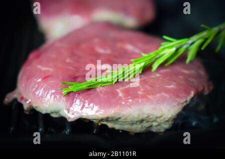 Leckeres Steak auf der Grillpfanne während des Garvorgangs; Nahansicht mit Fokus auf Rosmarinsprig Stockfoto