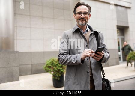 Bild eines fröhlichen Geschäftsmannes in Brillen mit Handy und Lächeln beim stehen in der Nähe des Bürogebäudes an der Stadtstraße Stockfoto