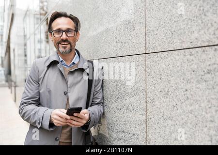 Bild eines freudigen erwachsenen Geschäftsmannes in Brillen mit Handy und Lachen, während er sich draußen an die Wand lehnt Stockfoto