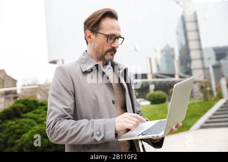 Bild eines konzentrierten Geschäftsmannes in Brillen, der Laptop verwendet, während er auf der Straße der Stadt steht Stockfoto