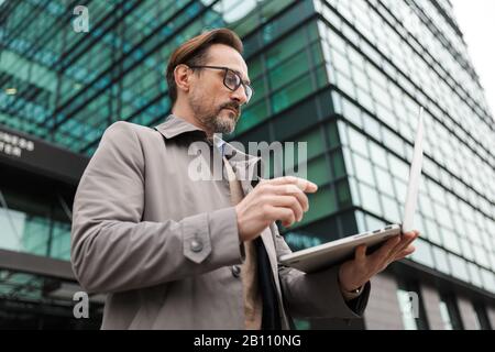 Bild eines reifen bärtigen Geschäftsmannes in Brillen, der Laptop verwendet, während er in der Nähe des Bürogebäudes in der Stadt steht Stockfoto