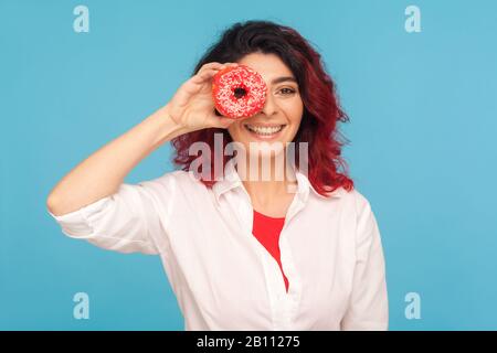 Süßes Süßigkeiten. Porträt der Hüpffrau mit ausgefallenen roten Haaren, die durch Donut und Lächeln schauen, Spaß mit Donut haben und köstliche Vergangenheit zeigen Stockfoto