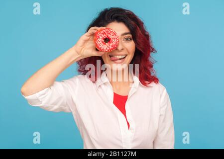 Süßer Snack, köstliches Gebäck. Porträt einer lächelnden Frau mit einem schicken roten Haar, das durch Donut schaut und die Zunge herausragt, Spaß mit Donut hat, Stockfoto