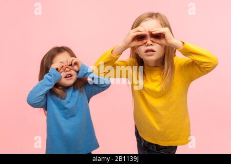 Liebenswürdige, neugierige, neugierige zwei kleine Mädchen, die durch die Finger schauen, das Fernglas imitieren, die Welt erkunden und etwas Neues mit interessiertem Blick entdecken Stockfoto