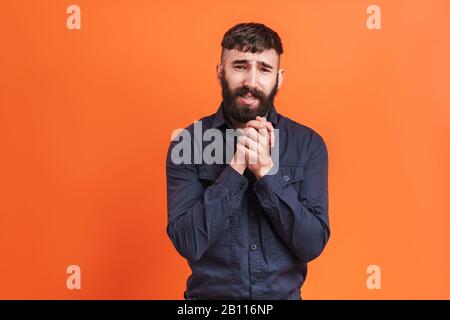 Bild Nahaufnahme des hochfesten Mannes mit Nasenschmuck, das schwarzes Hemd trägt und die Hände isoliert über orangefarbenem Hintergrund zusammenhält Stockfoto