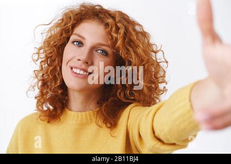 Nahaufnahme attraktive, fröhliche lächelnde Rotkopffrau mit lockigen Haaren, die die Hand nach vorne ziehen, als sie das Smartphone hält, selfie auf die Kamera nimmt, grinst Stockfoto