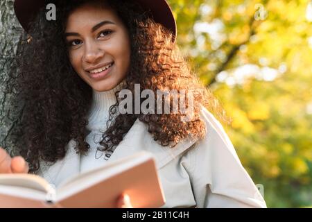 Schöne lächelnde afrikanerin, die einen Herbstmantel trägt, der sich an einen Baum lehnt, während sie im Park steht Stockfoto