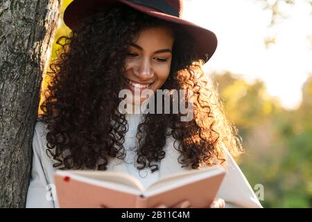 Schöne lächelnde afrikanerin, die einen Herbstmantel trägt, der sich an einen Baum lehnt, während sie im Park steht Stockfoto