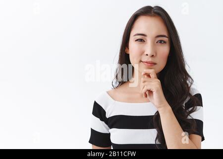 Skeptische junge asianerin im gestreiften T-Shirt, berühre das Kinn nachdenklich, schwelge begeistert von dem, was sie sieht, schlingt, schaue etwas mit Judgmentum an Stockfoto