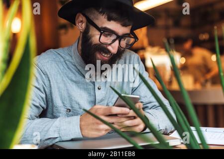Foto des freudigen jungen Mannes, der Brille mit Handy trägt und lächelt, während er im Café drinnen am Tisch sitzt Stockfoto