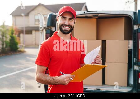 Bild des fröhlichen Lieferers in roter Uniform mit Zwischenablage und Schreiben in der Nähe des Autos mit Paketboxen im Freien Stockfoto