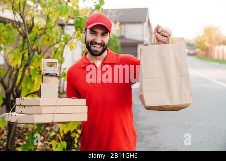 Bild eines lächelnden jungen Lieferers in roter Uniform, der Pizzakisten und Papiertüten auf der Straße in der Stadt hält Stockfoto