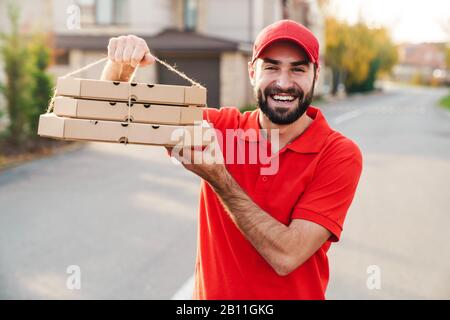 Bild des freudigen jungen Lieferers in roter Uniform, der Pizzakisten hält und auf der Straße in der Stadt lächelt Stockfoto