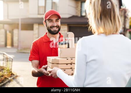 Bild eines lächelnden Auslieferers in roter Uniform, der der kaukasischen Frau auf der Straße der Stadt Speisenbestellung gibt Stockfoto