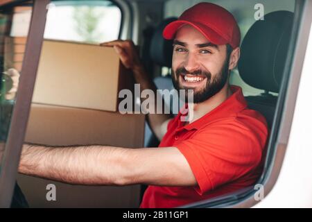 Bild eines fröhlichen jungen Lieferers in roter Uniform lächelnd und fahrender Lieferwagen mit Paketboxen Stockfoto