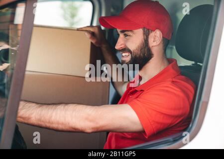 Bild eines fröhlichen jungen Lieferers in roter Uniform lächelnd und fahrender Lieferwagen mit Paketboxen Stockfoto