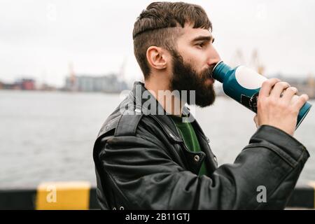 Foto eines bärtigen jungen Mannes, der Lederjacke trägt, die aus der Flasche trinkt, während er im Hafen steht Stockfoto