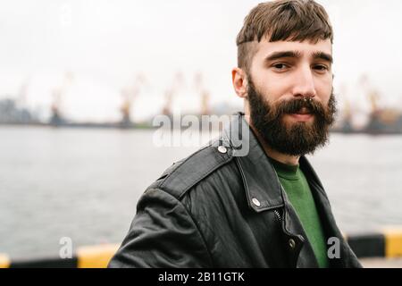 Fotoaufnahmen von bärtigen jungen Männern, die Lederjacke tragen und dabei auf die Kamera blicken, während sie im Hafen stehen Stockfoto