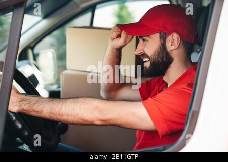 Bild eines fröhlichen bärtigen Lieferers in roter Uniform lächelnd und fahrender Lieferwagen mit Paketboxen Stockfoto