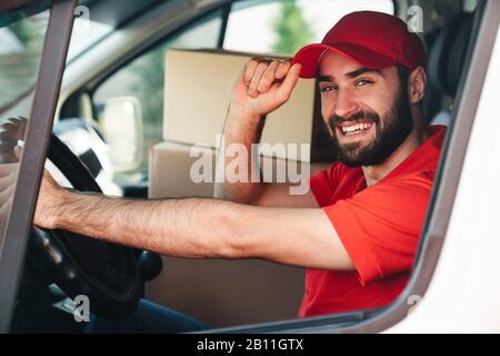 Bild eines fröhlichen bärtigen Lieferers in roter Uniform lächelnd und fahrender Lieferwagen mit Paketboxen Stockfoto