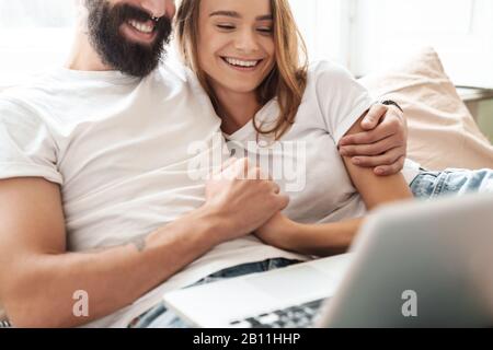 Das Bild eines kaukasischen Lachpaares mit Laptop und Umarmung im hellen Zimmer zu Hause Stockfoto
