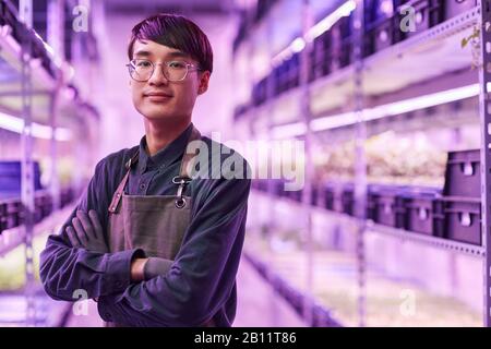 Porträt des asiatischen Jungbauers in der Schürze, die mit gekreuzten Armen steht und während der Arbeit auf der Farm die Kamera betrachtet Stockfoto