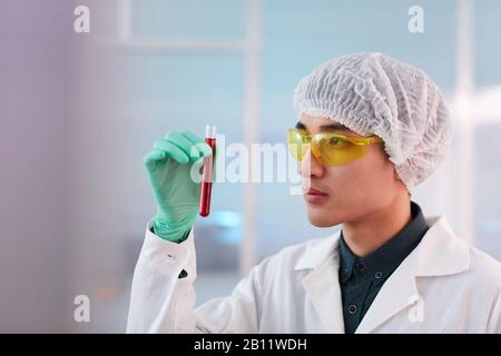 Asiatischer junger Wissenschaftler in schützenden Brillen und Verschlusskappe, die das Reagenzglas mit Blut hält Stockfoto