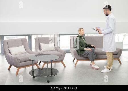 Junger Arzt in Whitecoat macht sich Notizen, während er von einer weiblichen Patientin steht Stockfoto