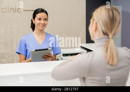 Fröhlicher junger Assistent, der vor dem Patienten an der Rezeption steht Stockfoto
