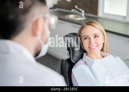 Fröhliche junge blonde Patientin, die ihren Zahnarzt mit gesundem Lächeln betrachtet Stockfoto