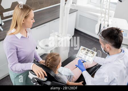 Junge Frau, die bei ihrer kleinen Tochter steht, die im Sessel des Zahnarztes sitzt Stockfoto