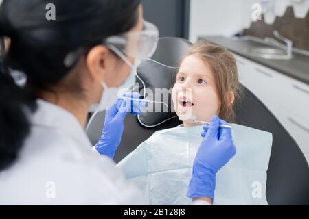 Kleines Mädchen mit offenem Mund, das den Zahnarzt in schützender Arbeitskleidung betrachtet Stockfoto
