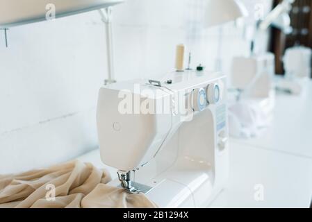 Elektrische weiße Nähmaschine mit beigefarbenem Stoff auf dem Schreibtisch in der Werkstatt Stockfoto