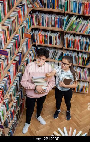 Hübsches Mädchen im Teenager-Alter in der Casualwear, das auf eines der Bücher im Bücherregal zeigt Stockfoto