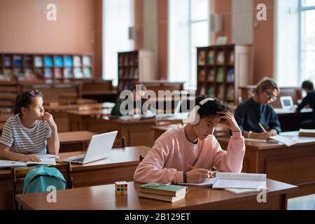 Gruppe zeitgenössischer Studenten, die an Schreibtischen in der Bibliothek sitzen Stockfoto