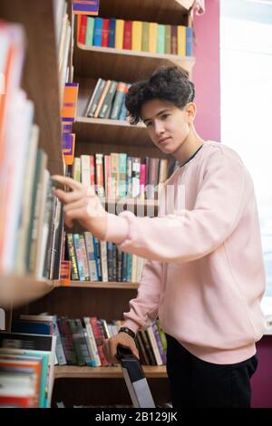 Ernster Teenager, der einen von Büchersaus großem Bücherregal in der College-Bibliothek mitnimmt Stockfoto