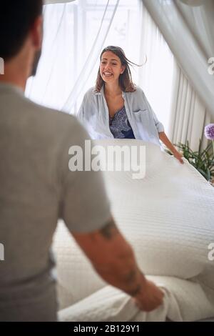 Das junge Paar spreizt mit Freude die Bettlaken im Schlafzimmer, lächelt: Schlafzimmer, Morgen, Routine, miteinander, Freude, Glückskonzept Stockfoto