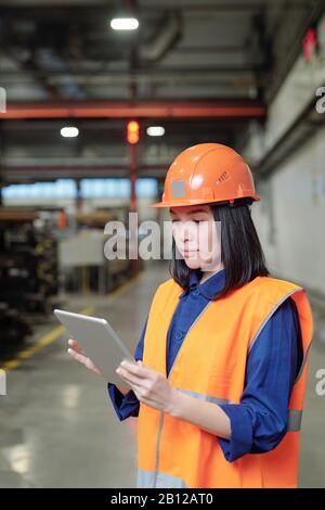 Ziemlich junge, ernsthafte Ingenieurin in Hardhat und Workwear mit Touchpad Stockfoto