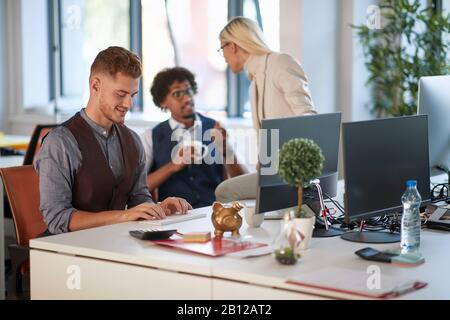 Weibliche Kollegin, die auf dem Schreibtisch sitzt und mit einer der Mitarbeiter spricht, während die dritte Person auf der Computertastatur tippt, zuhört. Business, Freizeit, Arbeit, Stockfoto