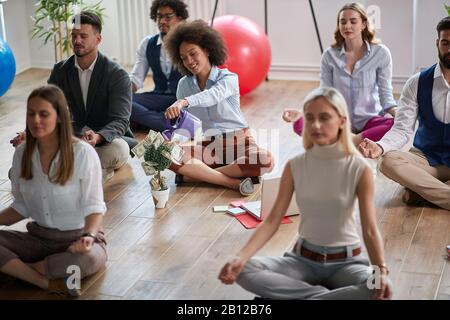 Eine Gruppe von Geschäftskollegen, die bei der Arbeit meditieren, auf dem Boden sitzen, eine Frau, die die Darstellung von Geldeinkommenssteigerung macht. Modern, Business, ich Stockfoto