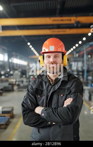 Fröhlicher junger bärtiger Industriearbeiter in Schutzkopfhörer und Helm Stockfoto