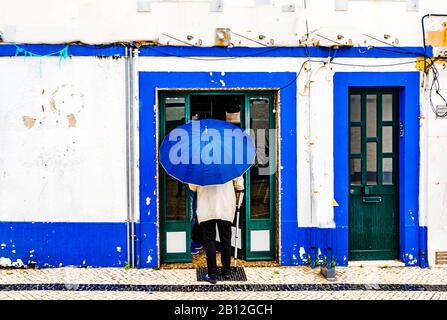 Blick auf Den Alten Mann mit Regenschirm vor dem blau gefärbten Gebäude in der Stadt Ericeira, Portugal Stockfoto