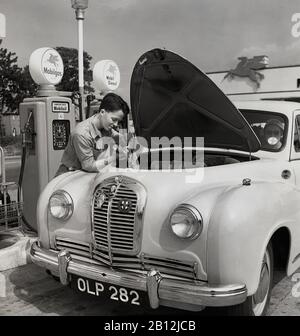 In den 1950er Jahren, historisch, an einer neuen 'Service Station, einem Auto der Ära, möglicherweise ein Austin, geparkt von den Kraftstoffpumpen mit offener Motorhaube und einem jungen männlichen uniformierten Garagenbegleiter, der den Ölmessstab oder die Öllehre des Motors überprüft, England, Großbritannien. Mit dem Anstieg des Autobesitzes in Großbritannien nach dem Krieg wurden größere Garagen oder "Servicestationen" eröffnet, die eine Reihe motorischer Dienstleistungen, einschließlich bemannter Vorfeldbegleiter, anbieten. Stockfoto