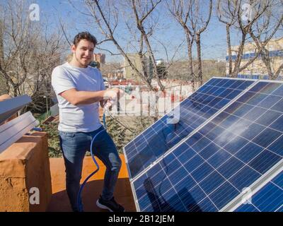 Kaukasischer junger Mann lächelt und säubert Solarpaneele mit einem Schlauch. Alternative Stromquelle, nachhaltige Ressourcenkonzeption. Stockfoto