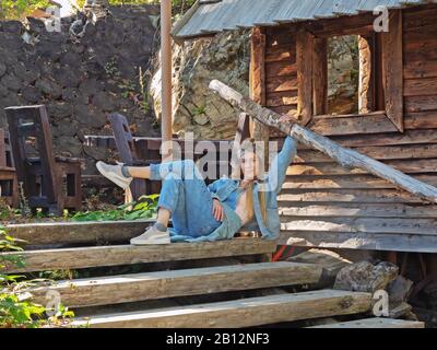 Sotschi, Russland - 13. Oktober 2019. Ein Mädchen in einem blauen Denimanzug sitzt auf Holztreppe, die am Geländer nahe der Holzhütte hält Stockfoto