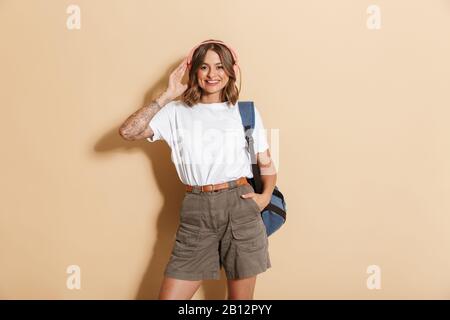 Porträt eines lächelnden Schulmädchens, das einen Rucksack trägt, der isoliert über beigefarbenen Hintergrund steht und Musik mit kabellosen Kopfhörern hört Stockfoto