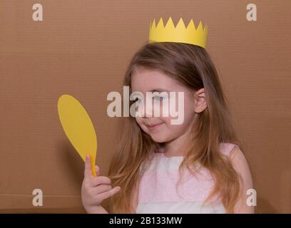 Portrait von emotionalen kleines Mädchen mit Krone ein gelbes Papier Holding ein Spiegel. handgefertigt. Karton Hintergrund. Vintage Style Stockfoto